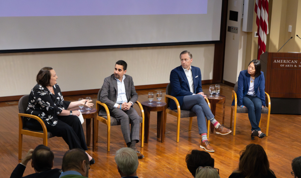 Four professionals participating in a panel discussion in front of an audience at an academic event, with logos of the hosting institutions displayed in the background.